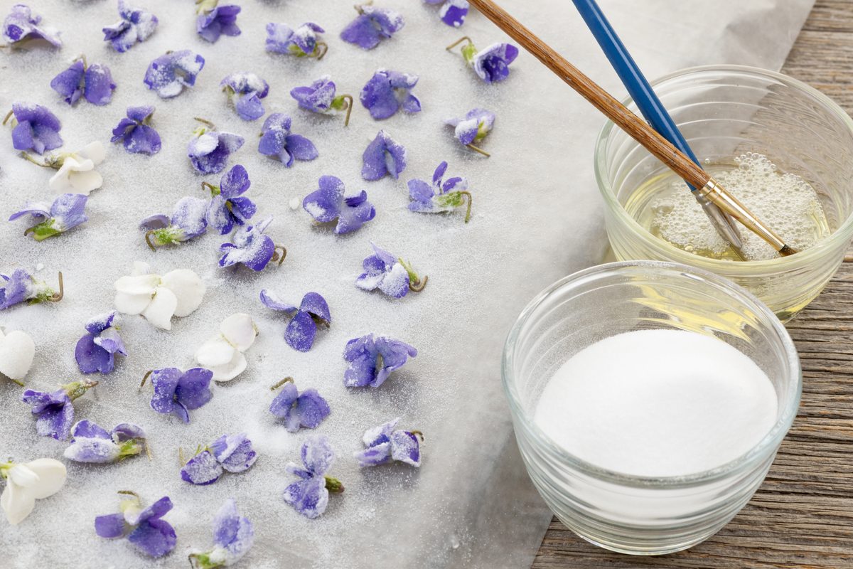 Candied violets on a baking sheet, with a small bowl of egg wash and a small bowl of sugar