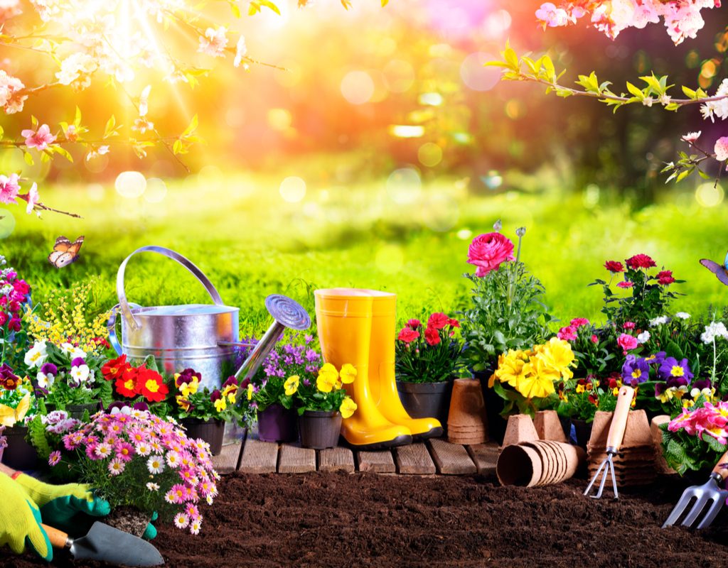 Yellow boots, metal garden tools and watering can, and assorted potted flowers in front of a patch of dirt. A gloved hand scoops some of the dirt in a little shovel.