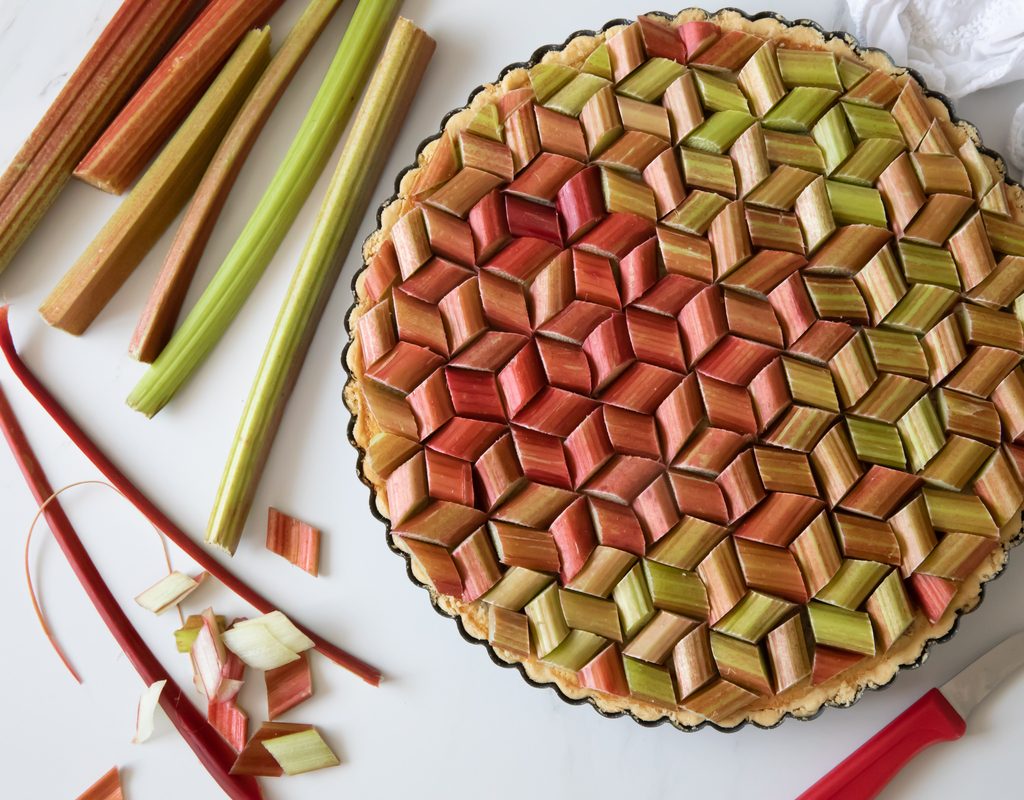 A fresh rhubarb pie with rhubarb pieces arranged geometrically on top and a few rhubarb stems sitting next to it
