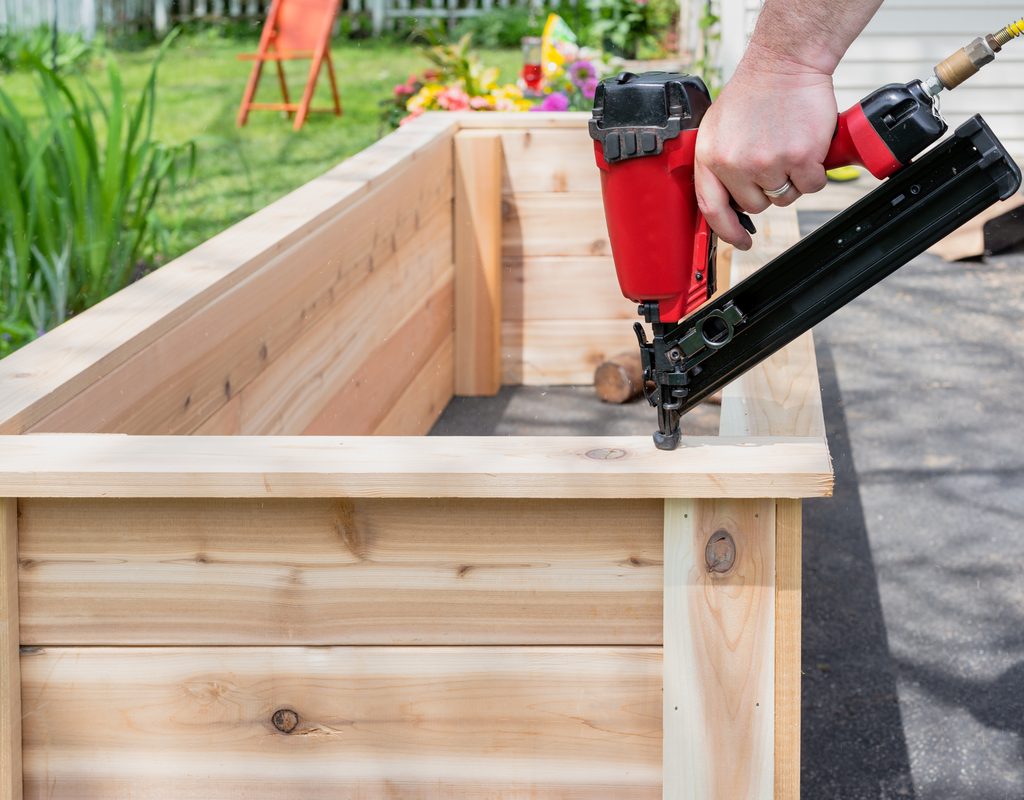 A person using a red nail gun to nail wood together, forming the frame of a raised garden bed