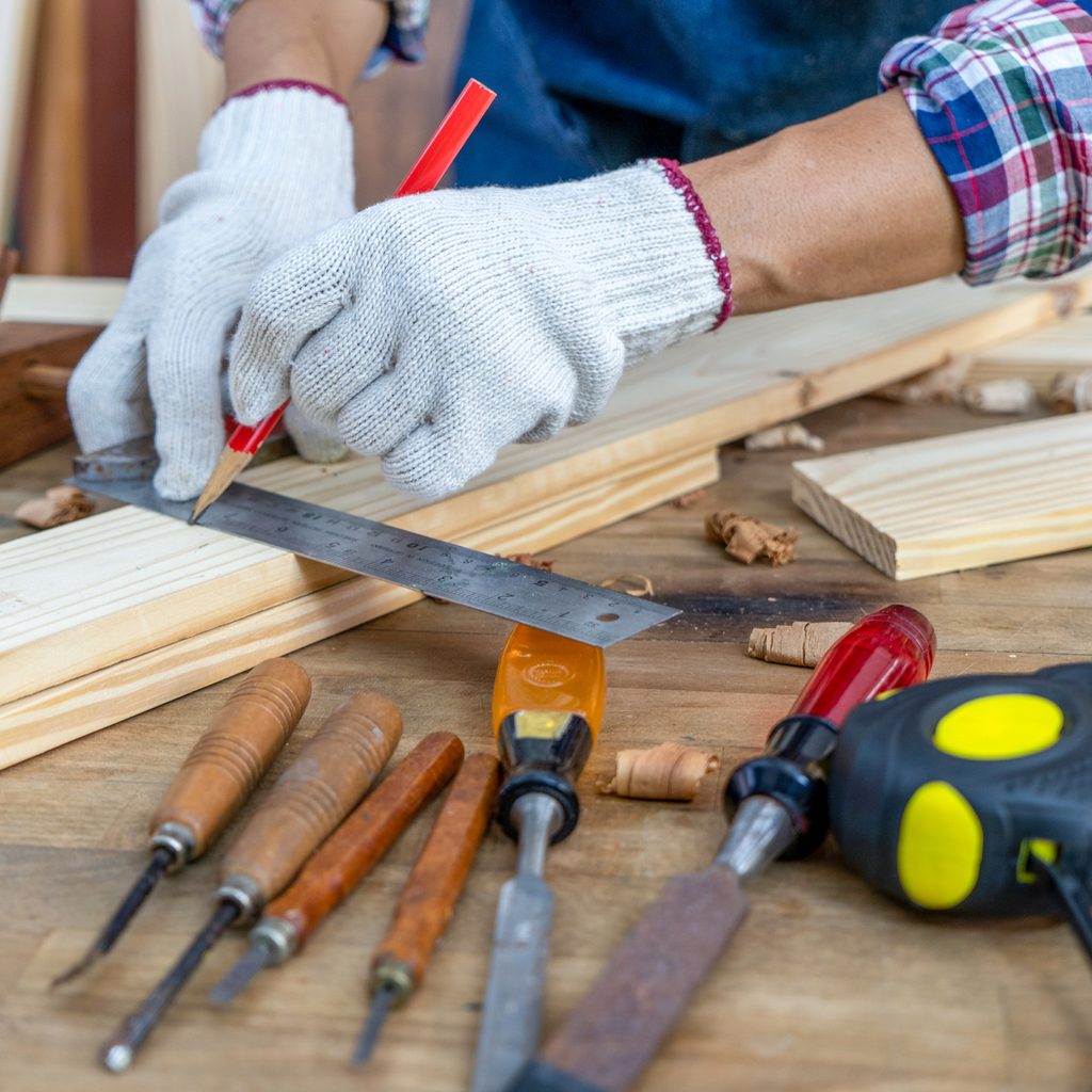 Aa person wearing work gloves, measuring a pieces of wood, surrounded by woodworking tools