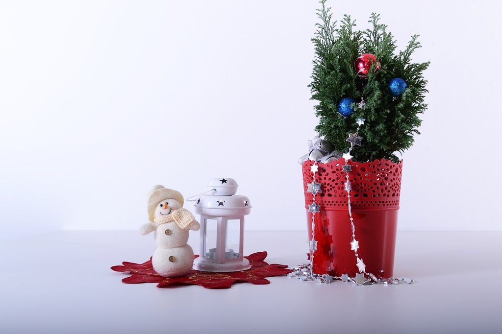 A small potted pine tree wrapped in Christmas decorations next to a tiny snowman figurine