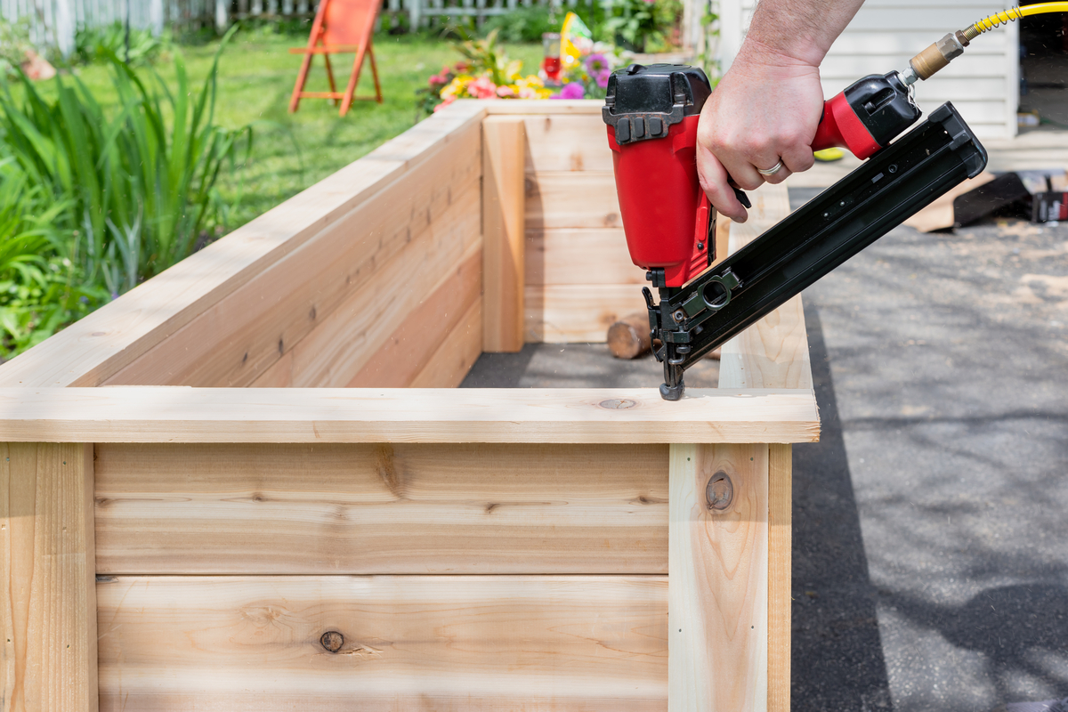 A person using a red nail gun to nail wood together, forming the frame of a raised garden bed