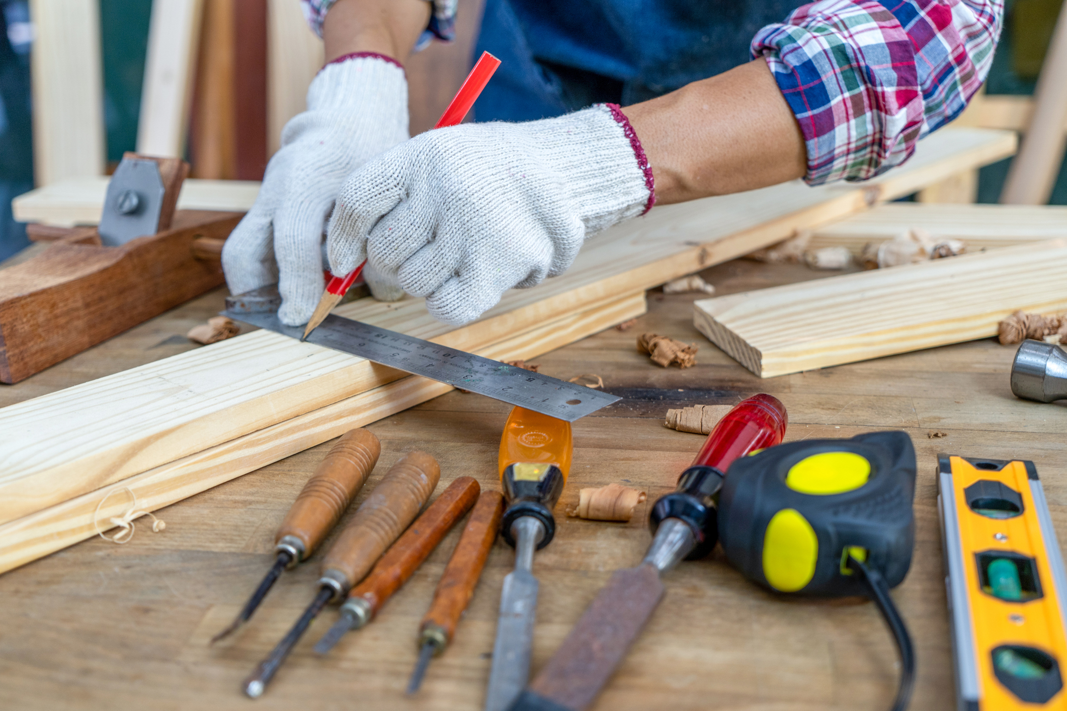 Aa person wearing work gloves, measuring a pieces of wood, surrounded by woodworking tools