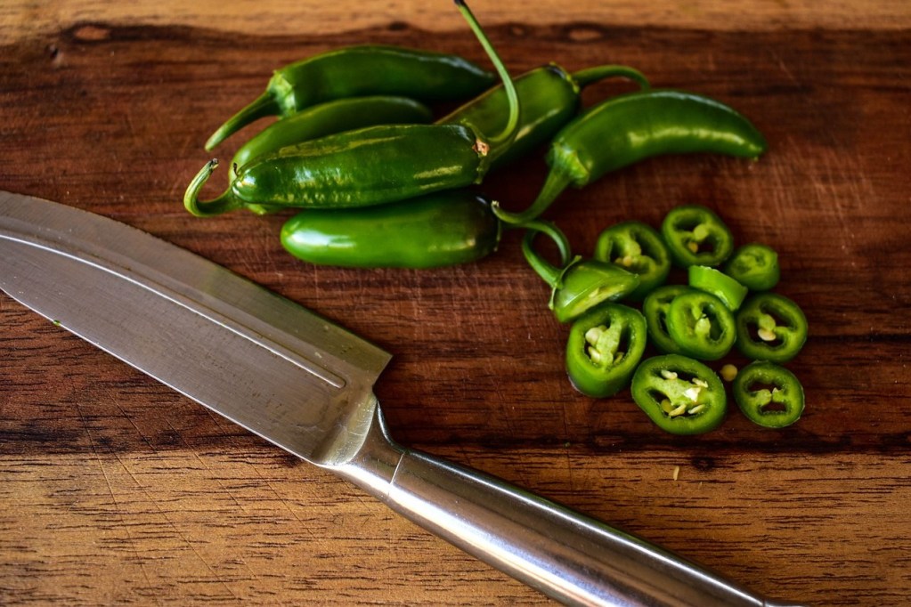 Several whole jalapenos next to a sliced jalapeno and a kitchen knife