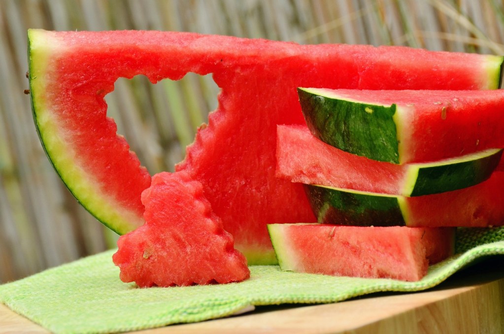 Watermelon slices in a stack. One slice has a heart-shape cut out of it, with the heart shaped watermelon piece propped in front of it.