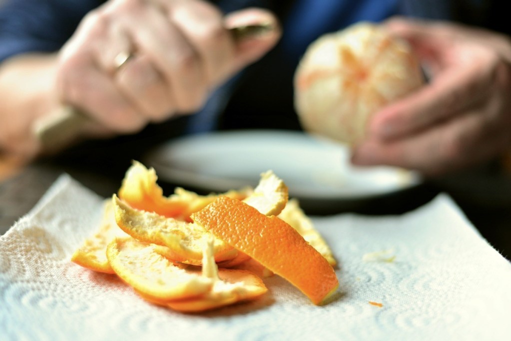 A pile of orange peels. In the background a man sits with a peeled orange and a knife