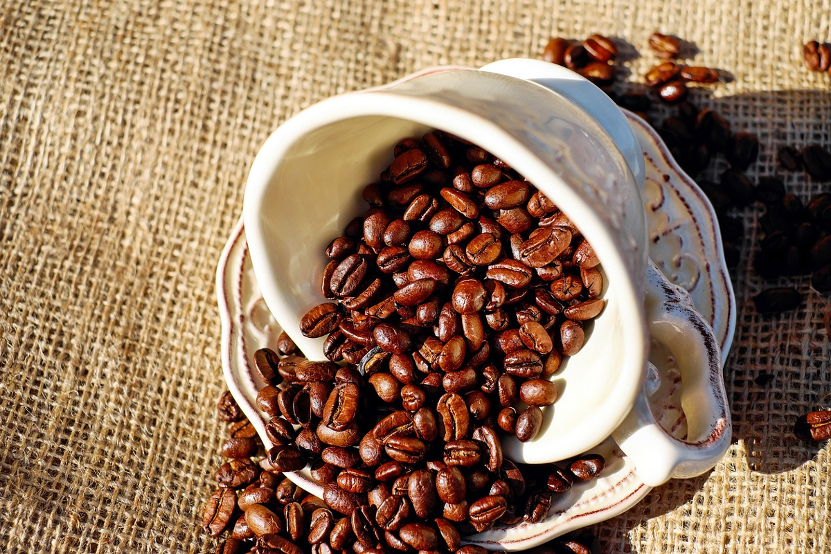 A white coffee cup tipped over on a matching saucer with roasted coffee beans spilling out of it